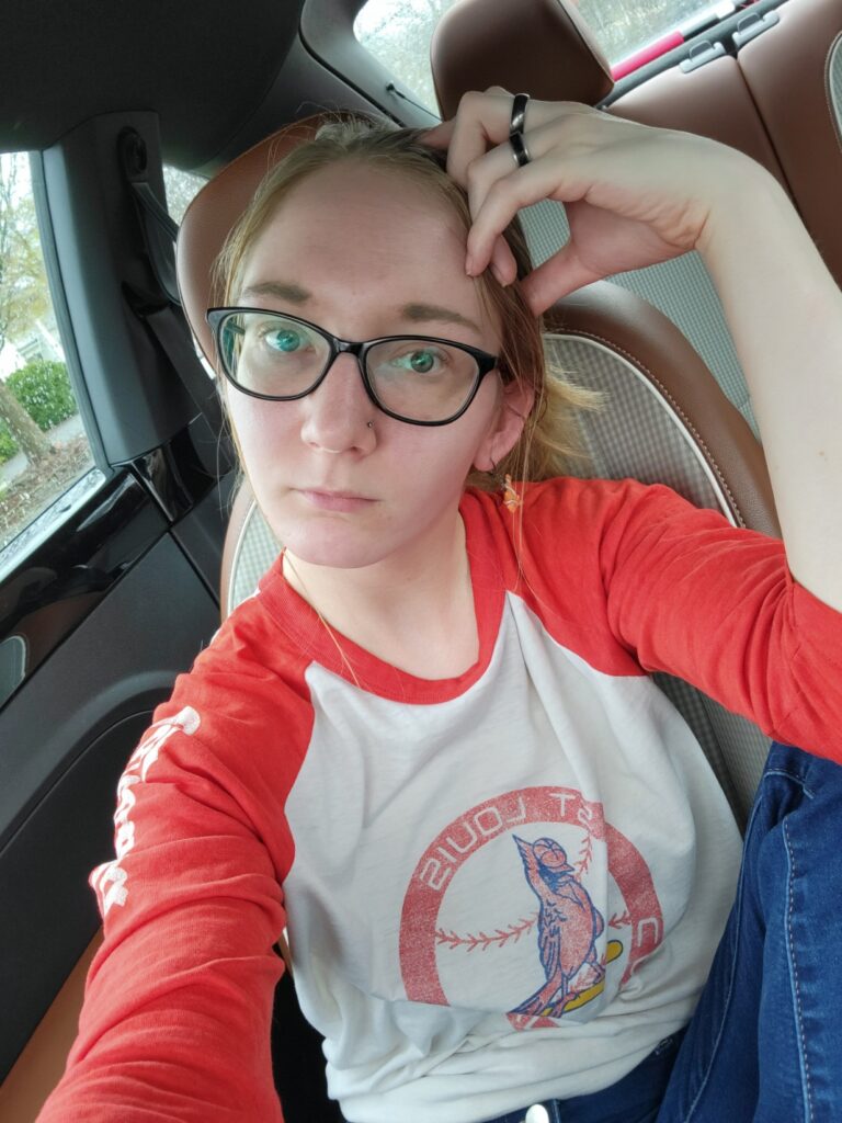 image of a small blonde with black glasses in a vintage Cardinals baseball tee inside a car thinking this will make her look cool.