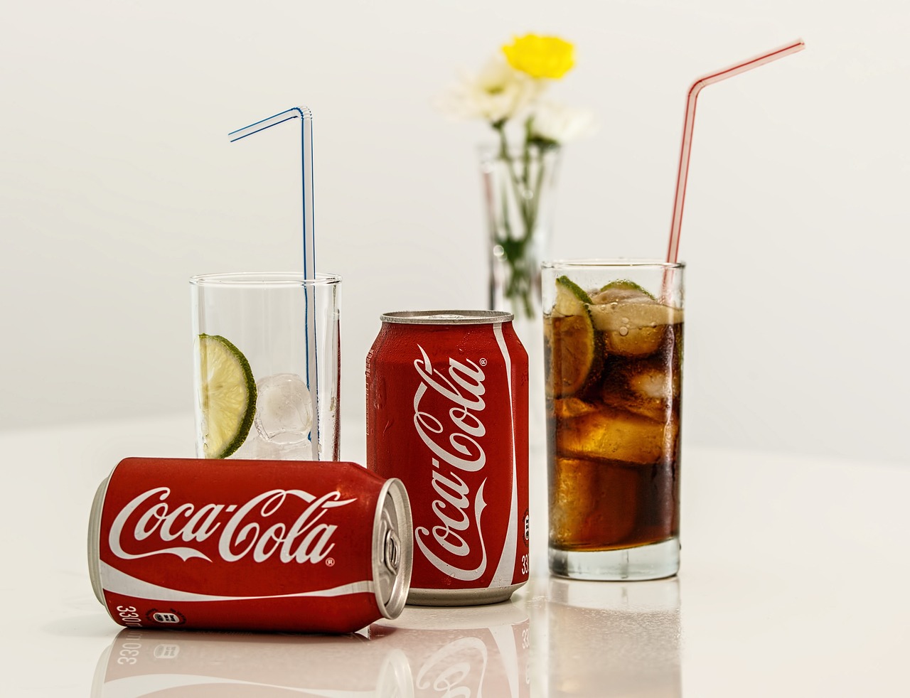 Coca cola can laying on side, two clear glasses with straws and one coca cola can standing right side up on a white table.